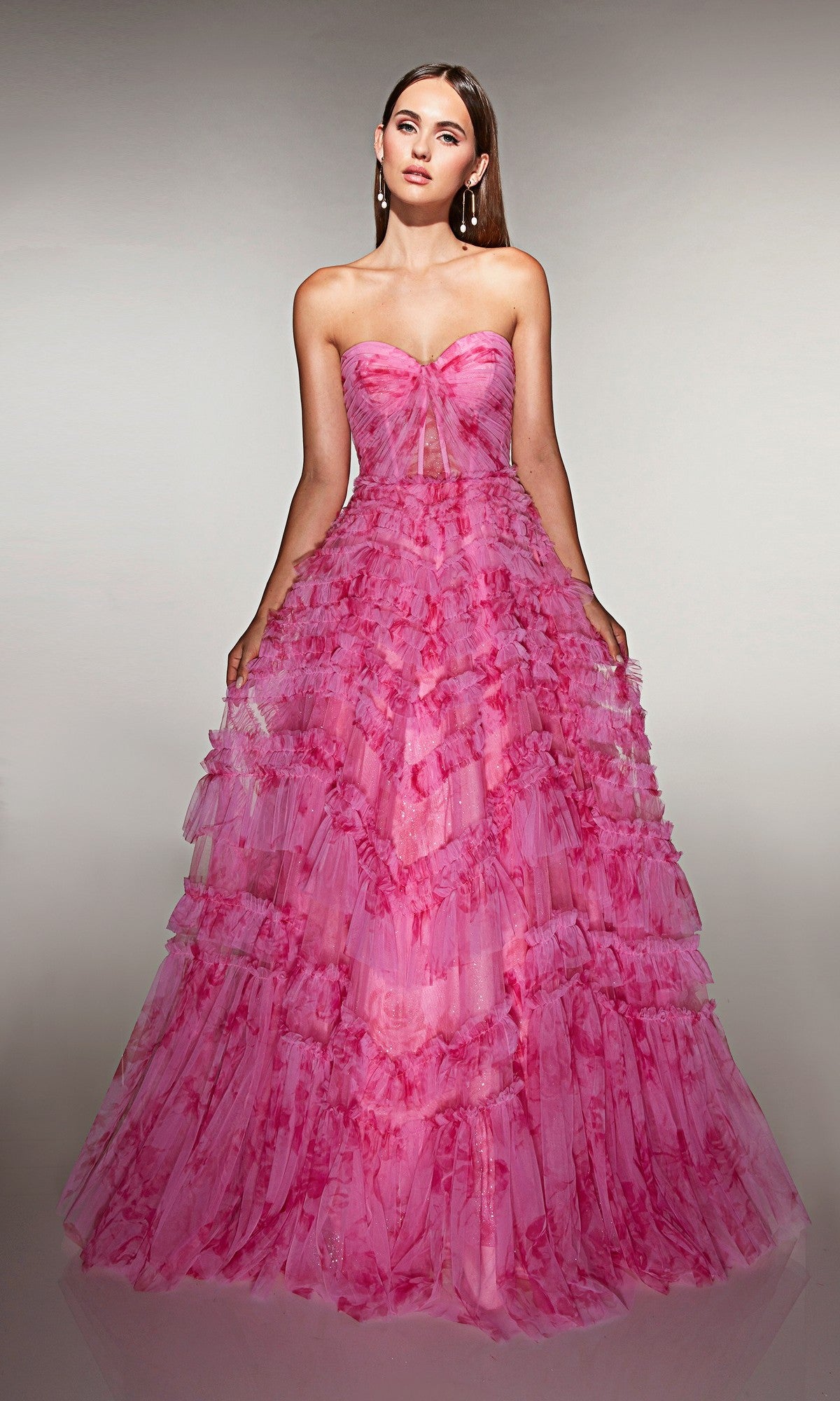 A woman wears the Alyce 62164 long prom dress, a strapless pink ballgown with a corset bodice and dramatic tiered ruffle skirt, standing against a neutral background.